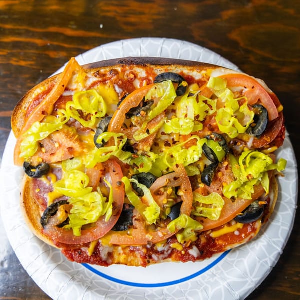 A toasted Sub Station pizza sub with pepperoni, tomatoes, olives, and banana peppers on a paper plate.