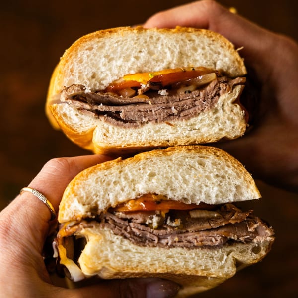 Hands holding a sliced roast beef sub sandwich with melted cheese and onions on a roll.